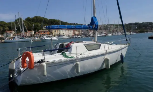 Bateau blanc à quai, voiles bleues enroulées. Au loin, un port animé avec des maisons colorées et d'autres voiliers. Un endroit idéal pour un séjour, comme le suggèrent les gib sea 76 avis.