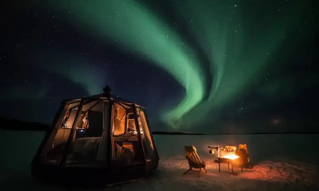 En glaskupol för camping i Norrland lyser upp natten under ett dansande norrsken. En eld sprakar vid sidan om.
