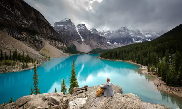Person sitzt auf Felsen und blickt auf den türkisfarbenen Moränen-See im Banff National Park, Kanada. Majestätische Berge und Nadelwälder umgeben das Wasser.
