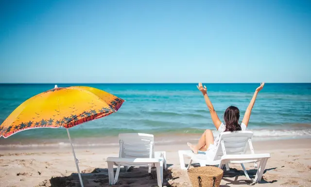 Kobieta cieszy się urlopem wypoczynkowym na plaży, z rękami w górze. Obok leżaki i parasol.