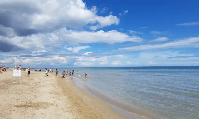 Una giornata di sole sulla spiaggia del cuore Riccione, con bambini che giocano e adulti che si godono il mare calmo sotto un cielo azzurro e nuvoloso.