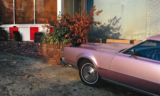 Un'auto rosa pastello, un classico stile William Eggleston, parcheggiata vicino a un muro di mattoni e un edificio con pannelli rossi e bianchi.