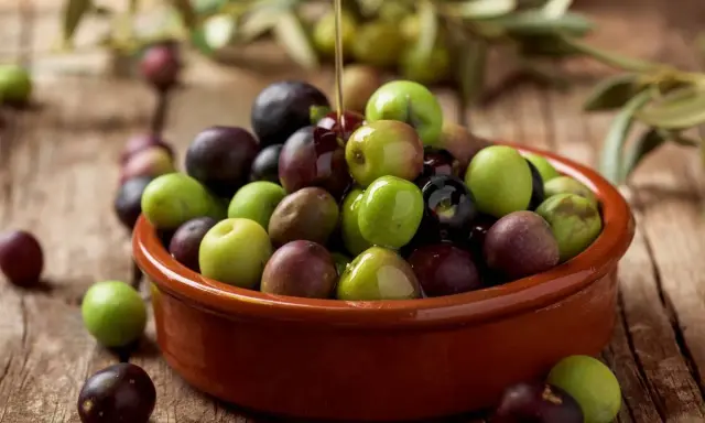 Aceite de arbequina vertiéndose sobre aceitunas verdes y moradas en un cuenco de barro. Ideal para ensaladas y aliños.