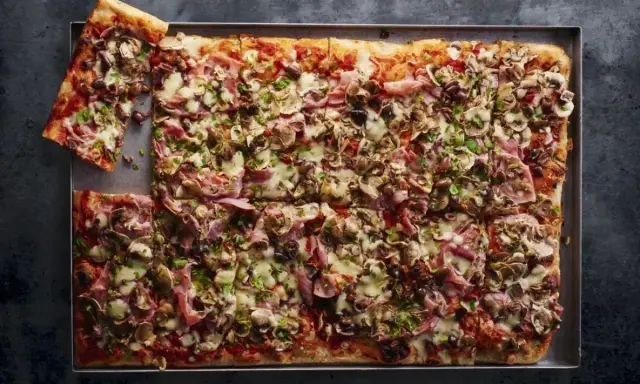 Prostokątna pizza 4 sery z pieczarkami, szynką i oliwkami, pokrojona na kwadraty, z jednym kawałkiem odłożonym na bok.