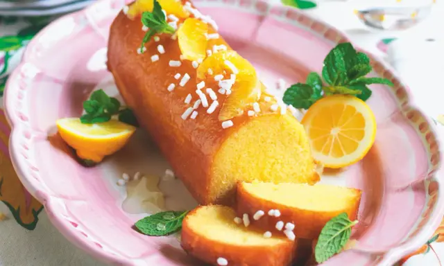 Deliziosa torta al limone glassata, decorata con fette d'arancia, zuccherini e menta. Servita su un piatto rosa con spicchi di limone.