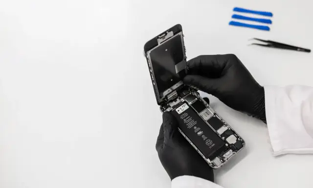 Ręce w czarnych rękawiczkach rozbierają iPhone'a, który wpadł do wody, ale działa. Widać baterię i wnętrze telefonu.