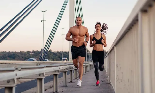 Ein Mann und eine Frau joggen auf einer Brücke. Beide haben sichtbare bauchmuskeln, ein Zeichen für ihr intensives Training.