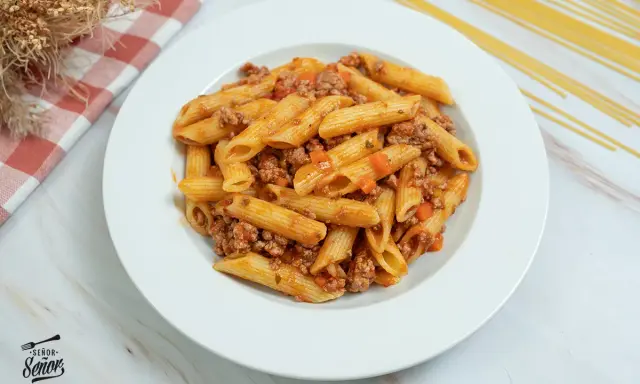 Plato de macarrones con carne picada y salsa de tomate, decorado con un paño a cuadros y espaguetis secos al fondo.