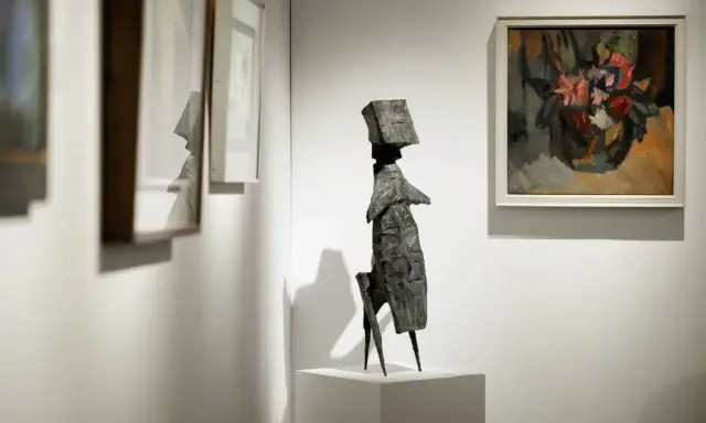 A striking bronze sculpture stands on a pedestal at the London Art Fair, surrounded by framed paintings.