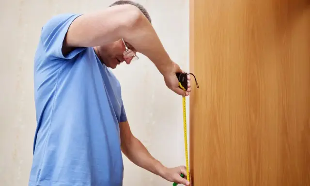 Mężczyzna w niebieskiej koszulce mierzy taśmą mierniczą drewniane drzwi, aby dowiedzieć się, jak zmierzyć drzwi zewnętrzne.
