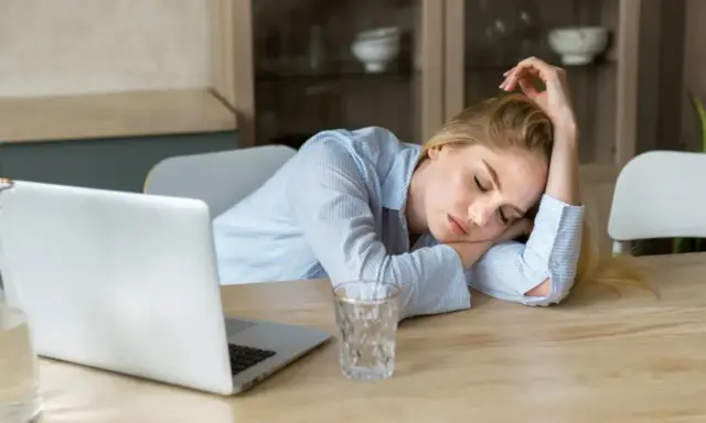 Zmęczona kobieta śpi na biurku obok laptopa. Objawy wypalenia zawodowego, czas na L4.