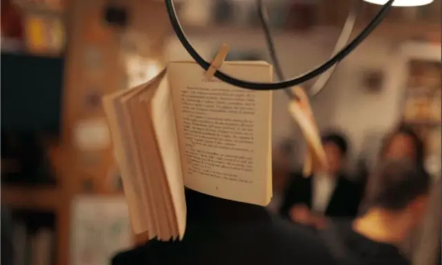 Un libro aperto, appeso per creare una scena da foto libri aesthetic. Le pagine sono piene di testo, con persone sfocate sullo sfondo.