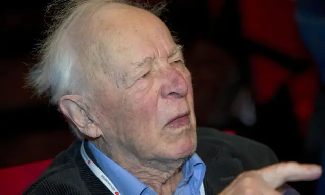 Äldre man med vitt hår, klädd i blå skjorta och mörk kavaj. Han bär ett band runt halsen med texten "Vänsterpartiet".
