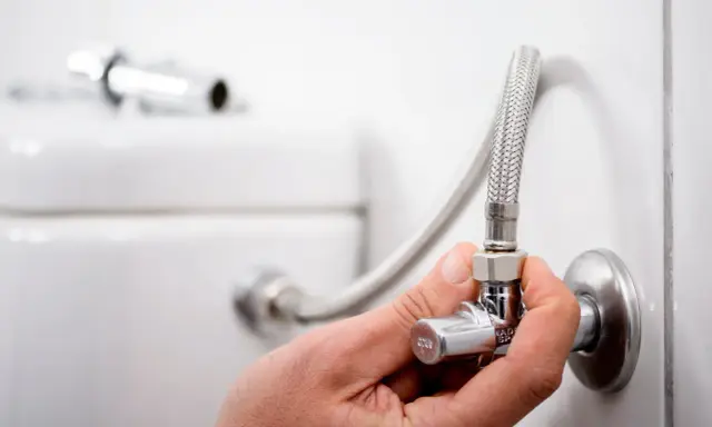 Mano ajustando la llave de paso del agua, que es comunitaria, conectada a una manguera flexible y al inodoro.