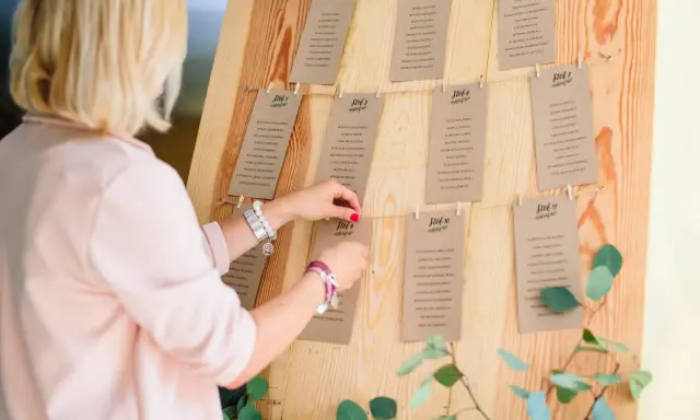 Ręcznie robiony plan stołów DIY na drewnianej tablicy, z kartkami przyczepionymi klamerkami. Kobieta poprawia jedną z kart.
