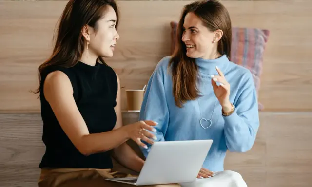 Dwie kobiety dyskutują przy laptopie. Jedna z nich, w czarnej bluzce, wskazuje coś na ekranie, druga, w błękitnym swetrze, słucha z uśmiechem, zastanawiając się, co to jest status.