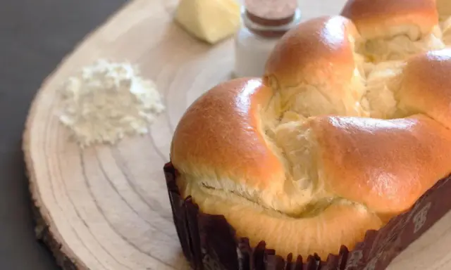 Un pan brioche dorado y esponjoso, listo para ser disfrutado. A su lado, ingredientes para su elaboración: harina, mantequilla y sal.