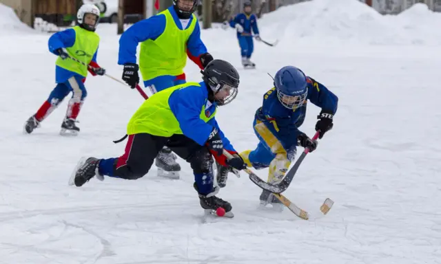 Barn i full fart med hockeyklubbor och hjälmar, ivriga att vinna pucken. De har roligt med lekar på isen.