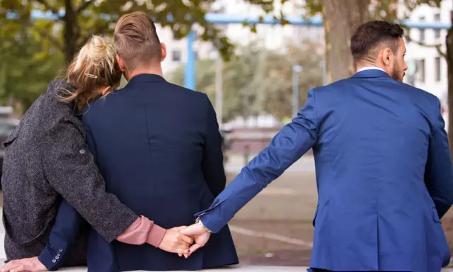 Trójkąt miłosny. Dwoje ludzi siedzi blisko siebie, a trzeci mężczyzna dyskretnie trzyma ich za rękę. To pokazuje, dlaczego zdrada emocjonalna tak boli.