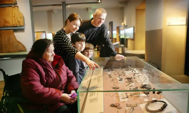 A family, including a woman in a wheelchair, examines artifacts in a museum display, perhaps inspired by famous art curators.