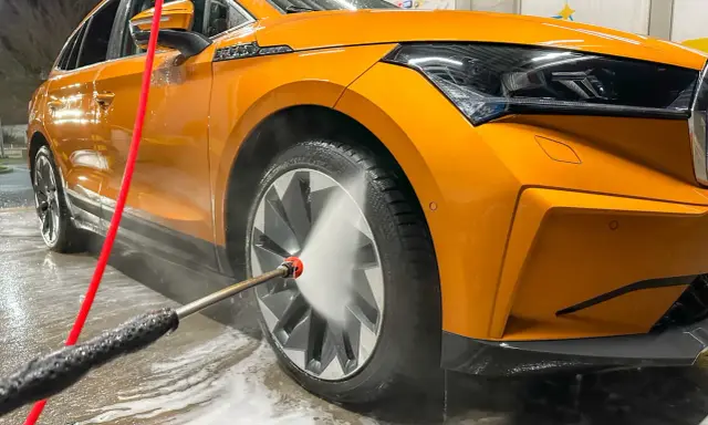 Pomarańczowe auto w trakcie mycia. Woda pod ciśnieniem spływa po karoserii i felgach, tworząc pianę.
