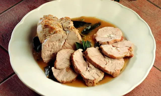 Delizioso arrosto di tacchino affettato, servito con salsa e foglie di salvia su un piatto bianco.