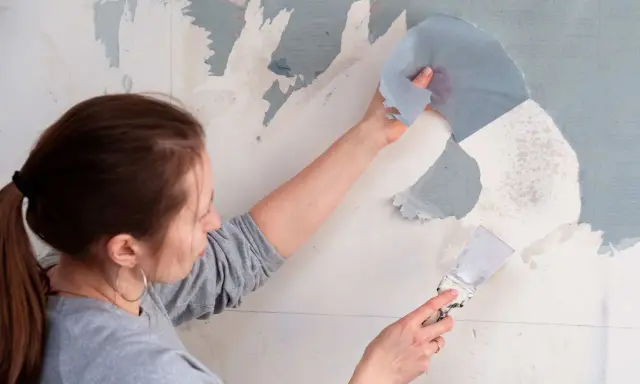 Une femme utilise une spatule pour décoller du papier peint ancien.