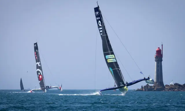 Course d'Ocean Fifty près d'un phare. Les trimarans filent sur l'eau, leurs voiles noires portant des logos.