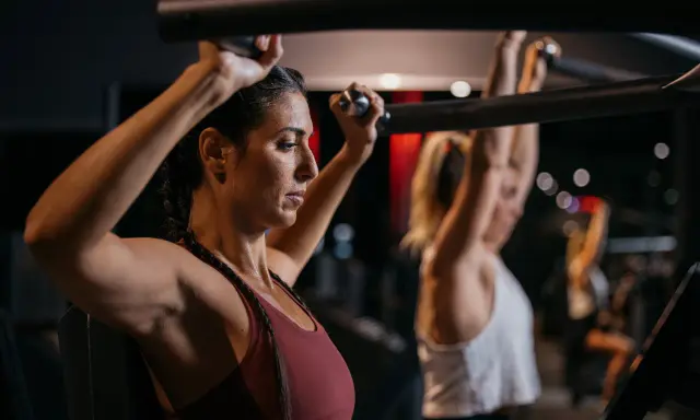 Zwei Frauen trainieren im Fitnessstudio. Sie nutzen eine Maschine, die zeigt, welche Muskelgruppen zusammen trainieren.