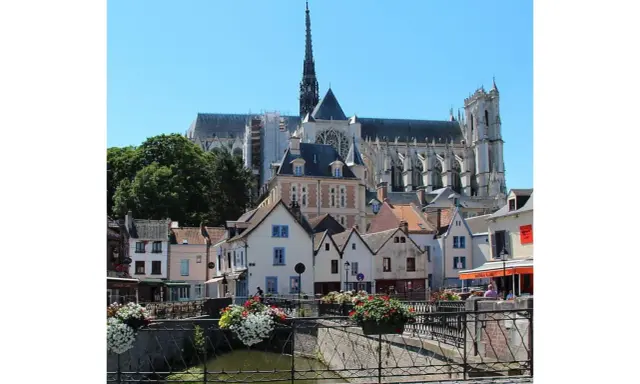 Blick auf malerische Häuser und die Kathedrale in Amiens, Frankreich. Blumenkästen schmücken eine Brücke über einen Kanal.