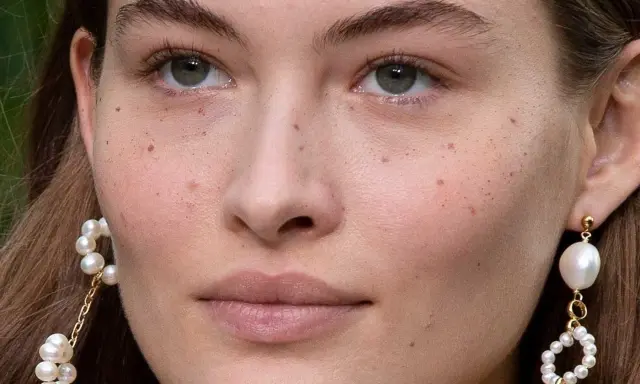 Primer plano de un rostro femenino con pecas naturales salpicando su piel. Lleva pendientes de perlas colgantes.