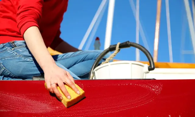 Maintenant, on apprend comment laver une coque de bateau. Une main savonneuse frotte la surface rouge avec une éponge jaune.