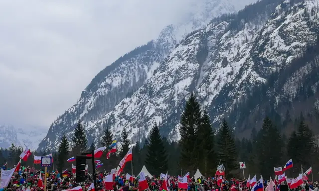 Tłum kibiców z flagami na tle ośnieżonej góry, atmosfera na skoczni w Planicy.