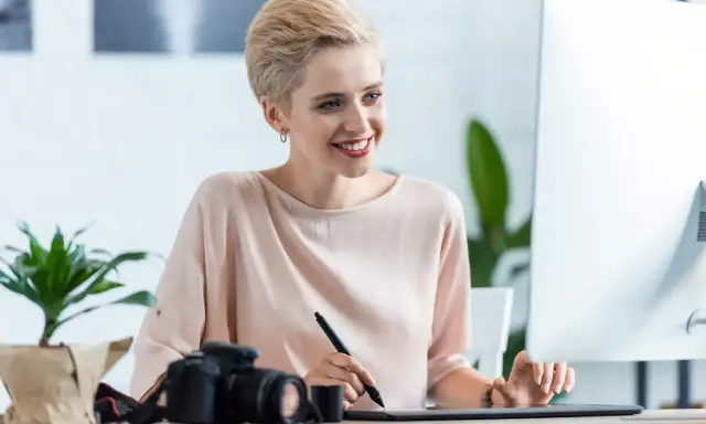 Uśmiechnięta kobieta z krótkimi blond włosami pracuje przy komputerze, obok aparat fotograficzny. Sprawdza najnowsze oferty pracy w Pile.