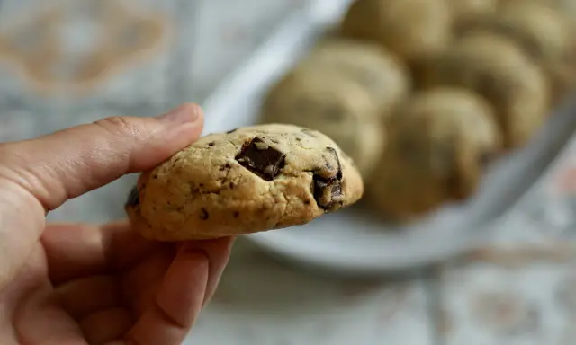 Une main tient un cookie épais et moelleux aux pépites de chocolat. D'autres cookies sont disposés sur une assiette en arrière-plan.