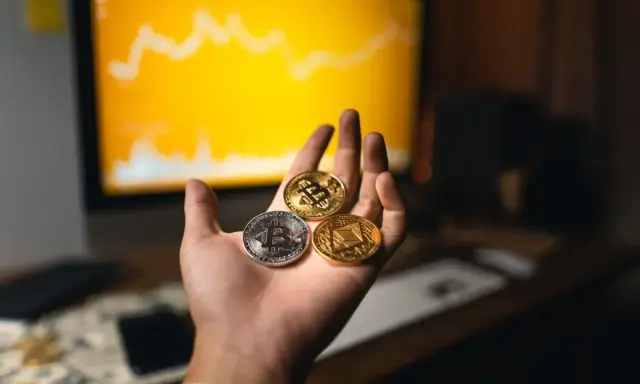 Dłoń trzyma trzy monety kryptowalut: Bitcoin, Ethereum i inną. W tle wykres na monitorze, sugerujący handel na kantory kryptowalut.