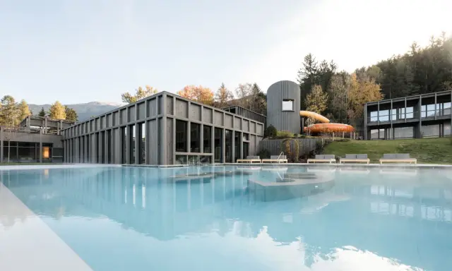 Piscina esterna con acqua fumante e scivoli acquatici arancioni al Aki Family Resort Plose. Architettura moderna in legno e montagne sullo sfondo.