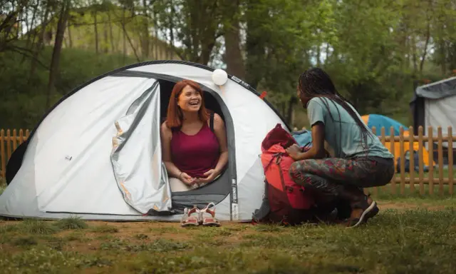 Dwie kobiety cieszą się biwakowaniem. Jedna siedzi w namiocie, druga obok niego, przy plecaku. Camping pole namiotowe to podstawa udanego wypoczynku.