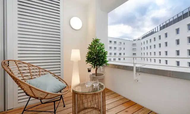 Przytulny balkon z drewna, z wiklinowym fotelem, stolikiem i rośliną. W tle białe budynki.