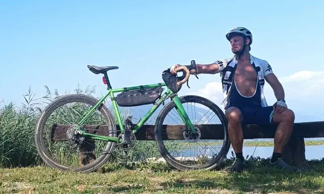 Ciclista riposa accanto alla sua bici verde, pensando alle tabelle per dimagrire in bicicletta. Cielo azzurro e lago sullo sfondo.