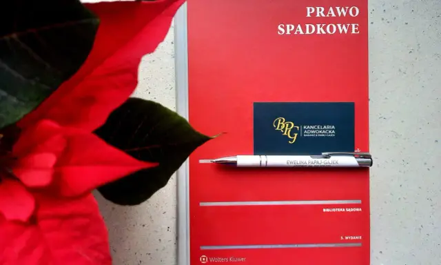 Czerwona książka "Prawo spadkowe" obok poinsecji. Długopis z nazwiskiem adwokatki i wizytówka kancelarii sugerują tematykę prawną, być może związaną z zapisem windykacyjnym.