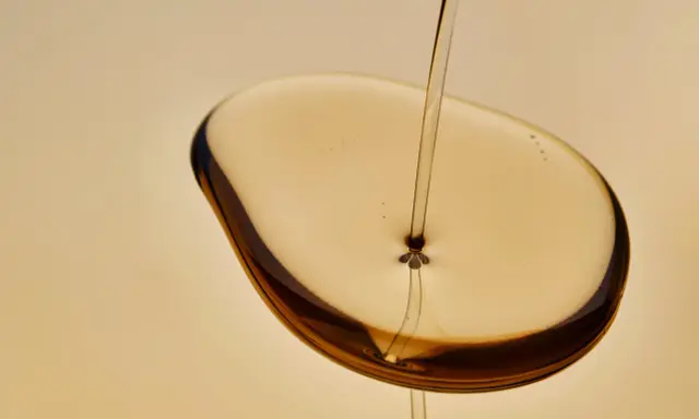 Un filet d'huile de karanja, d'une couleur ambrée, tombe sur une surface liquide, créant des reflets sombres. Attention, l'huile de karanja peut présenter un danger.