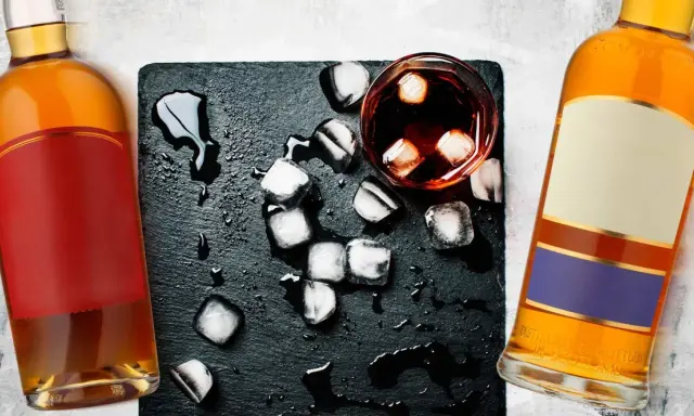 Dwie butelki, jedna z czerwoną etykietą, druga z beżową i fioletową, stoją obok szklanki z lodem i ciemnym płynem. Koniak czy whisky?
