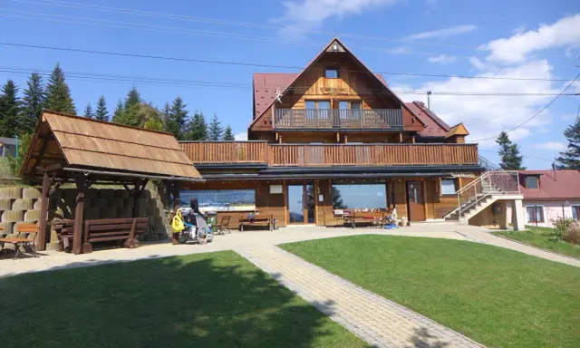 Drewniana bacówka na obidzy z balkonem i tarasem, otoczona zielenią i ścieżkami. Obok wózek inwalidzki z żółtą torbą.