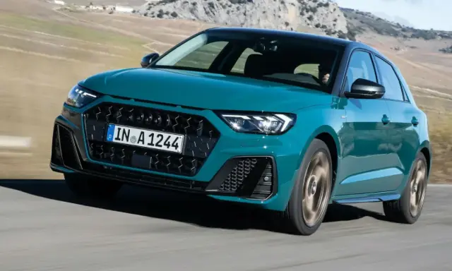Audi RS1 verde azulado en movimiento por una carretera rural con montañas al fondo.