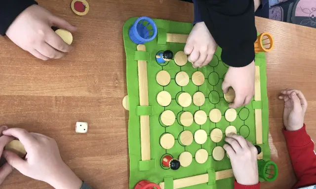 Dzieci grają w grę planszową podczas zajęć korekcyjno kompensacyjnych. Na zielonej macie są drewniane pionki i kostka.