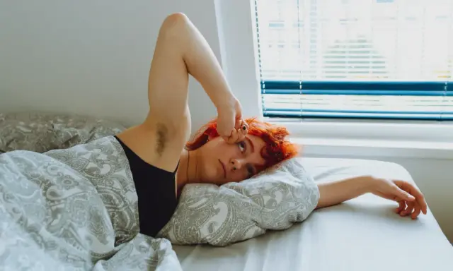Jeune femme aux cheveux roux allongée dans son lit, le bras levé, le regard perdu. Elle semble fatiguée, peut-être un des symptômes manque magnésium.
