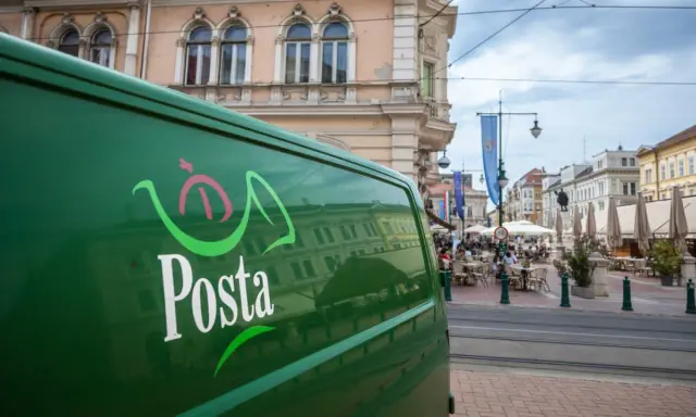 Zielona furgonetka pocztowa z logo "Posta" stoi na ulicy w węgierskim mieście. W tle widać kawiarnie i zabytkowe budynki.
