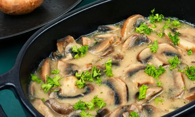 Pyszne pieczarki w śmietanie, doprawione świeżą natką pietruszki, podane w czarnej, owalnej misie.