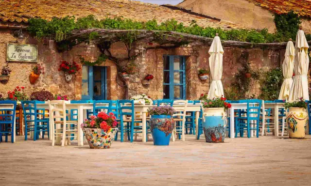 Taverna La Cialoma, un angolo incantevole di Siracusa e dintorni, con tavoli apparecchiati e sedie blu sotto un pergolato fiorito.